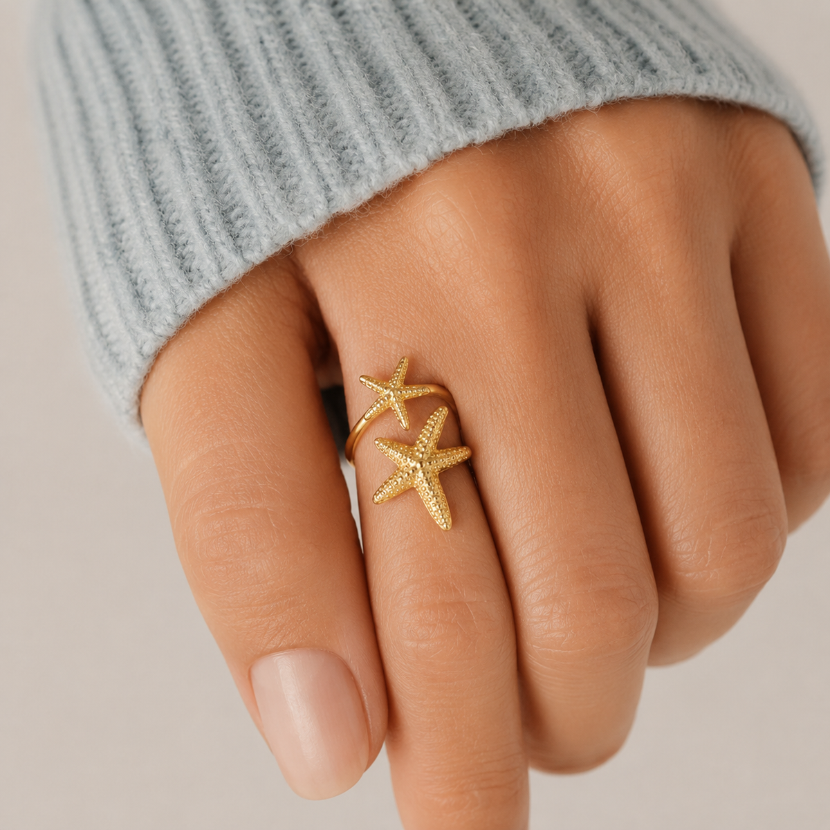 Anillo ajustable dorado con dos estrellas de mar texturizadas llevado en el dedo, combinado con un jersey azul claro para un look elegante, femenino y sofisticado.