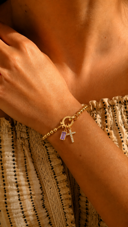 Pulsera dorada con cuentas y colgantes de cruz engastada y piedra amatista rectangular, destacada en la muñeca bajo luz cálida.