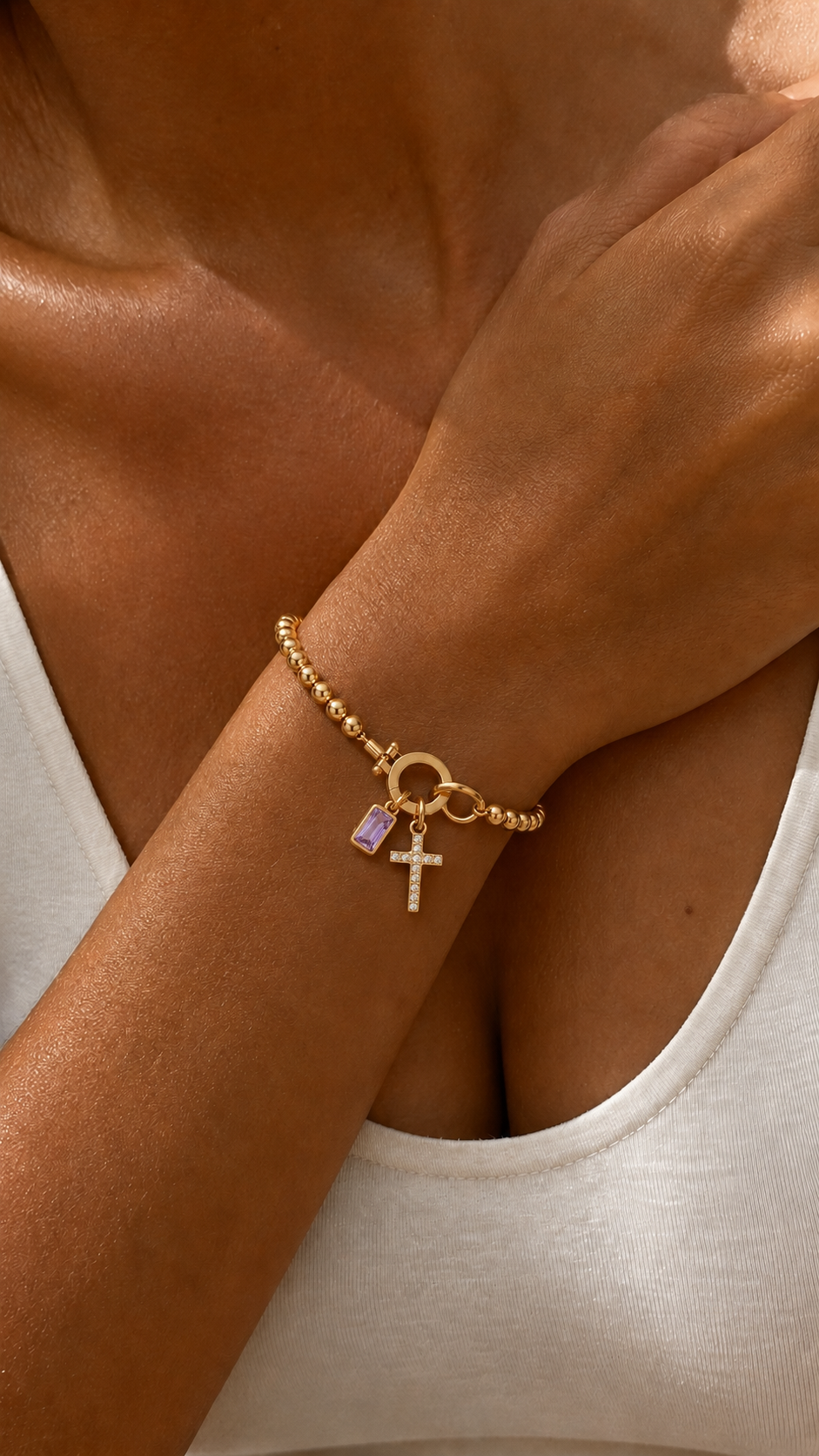 Pulsera dorada con cuentas y colgantes de cruz engastada y piedra amatista rectangular, destacada en la muñeca bajo luz cálida.