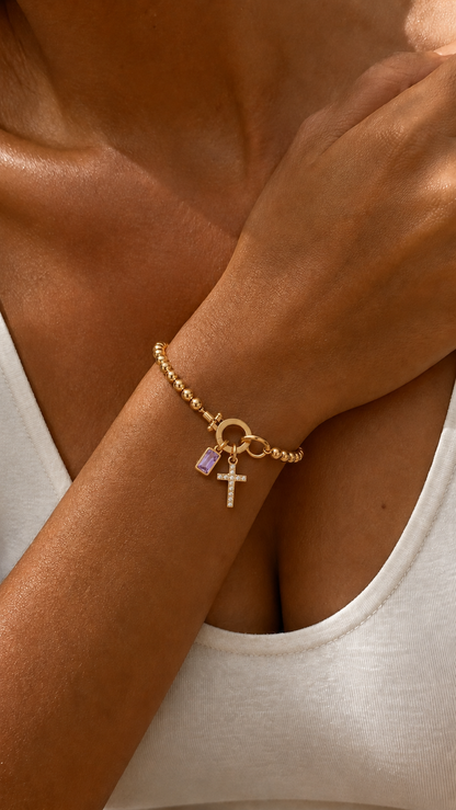 Pulsera dorada con cuentas y colgantes de cruz engastada y piedra amatista rectangular, destacada en la muñeca bajo luz cálida.