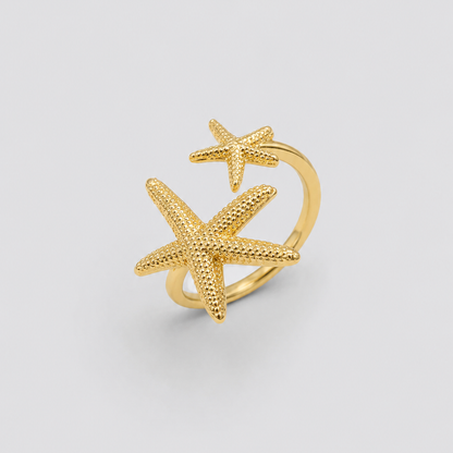 Anillo ajustable dorado con dos estrellas de mar texturizadas en tamaños diferentes, fotografiado sobre fondo gris claro para un look elegante, minimalista y sofisticado.
