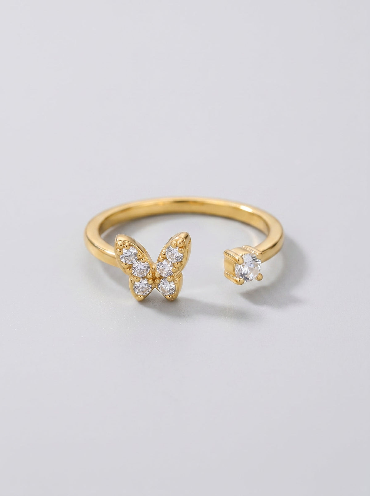 Anillo mariposa dorado con piedras preciosas transparentes sobre fondo gris claro.