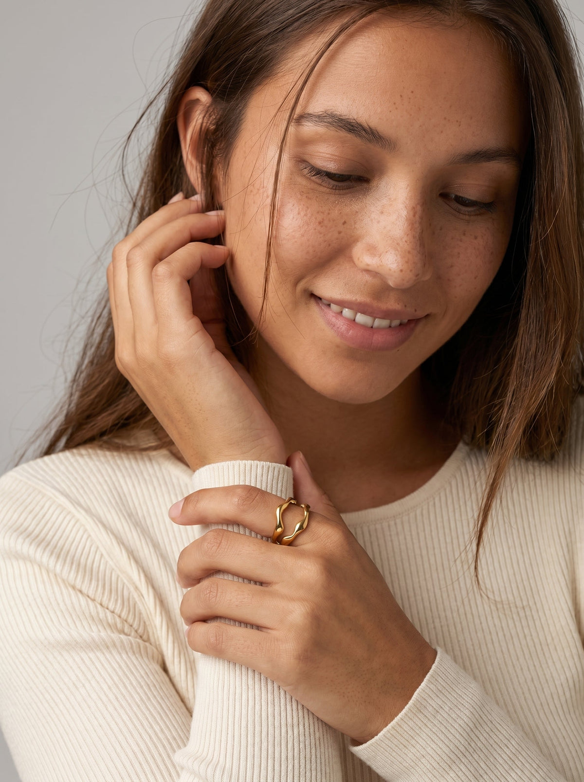 Mujer con un anillo dorado en el dedo, tocándose la oreja sobre un fondo neutro.
