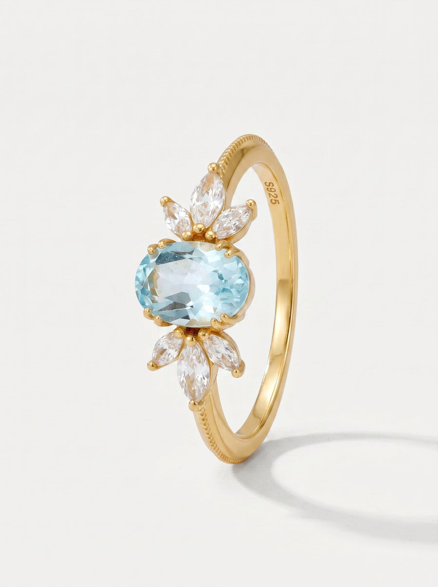 Anillo dorado con una piedra preciosa azul y detalles de diamante sobre un fondo gris claro.