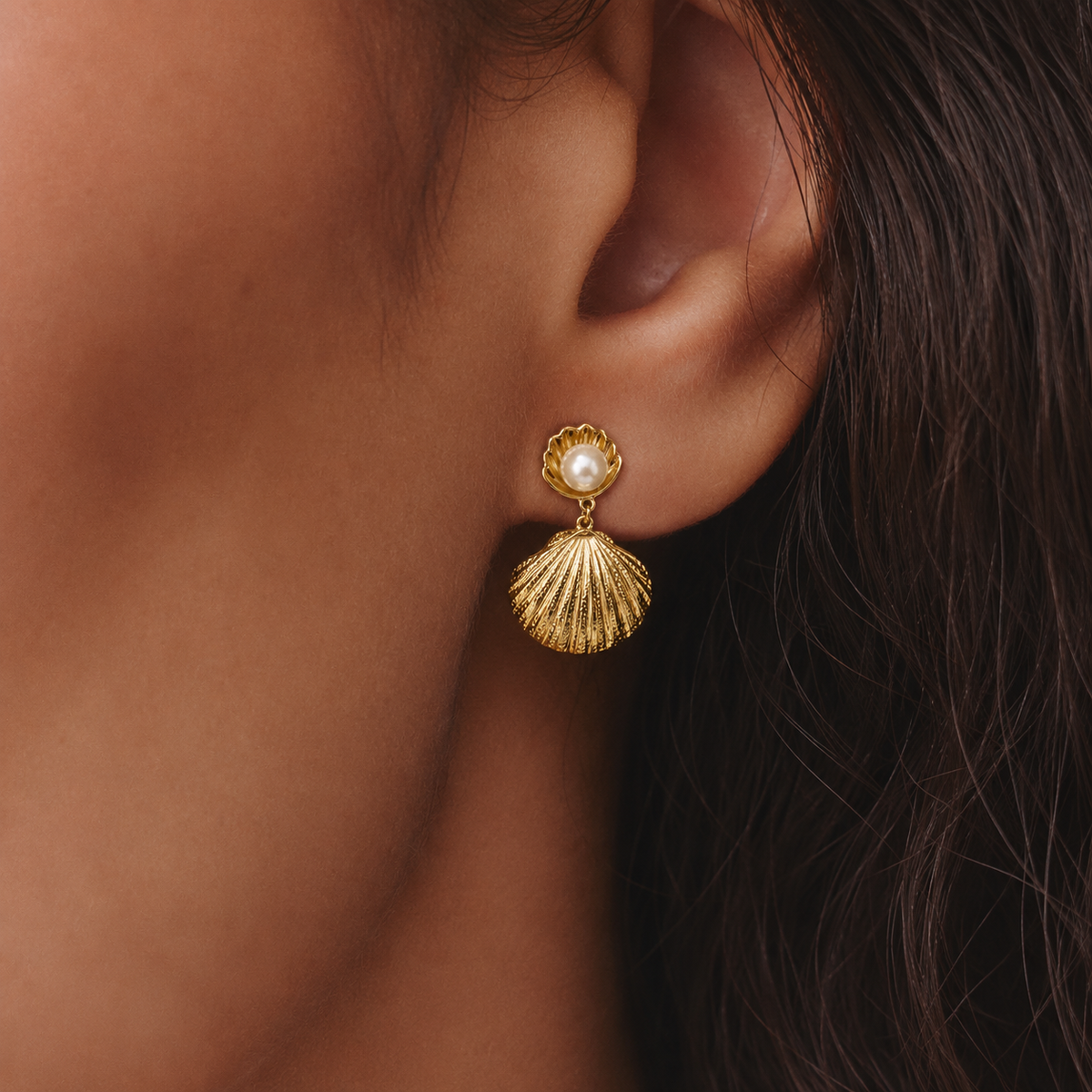 Pendientes dorados con concha y perla blanca llevados en la oreja, para un look elegante, femenino y sofisticado.