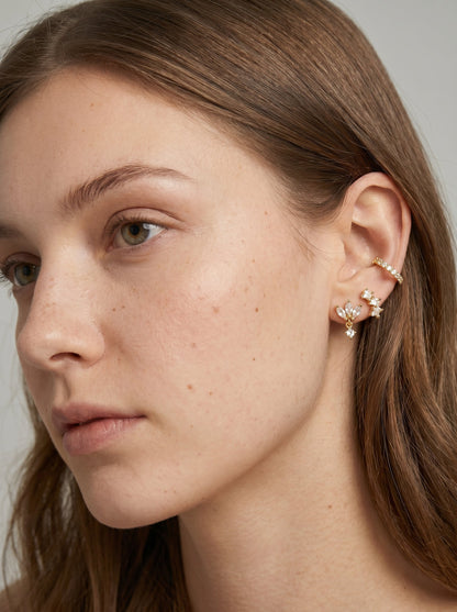 Pendientes de presión dorados y delicados pendientes florales con detalles en cristal transparente, mostrados en una modelo para un look elegante de joyería.

