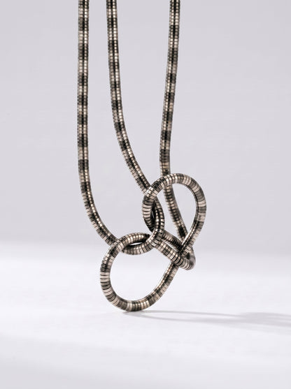 Collar plateado moderno con colgante abstracto en forma de nudo y acabado texturizado, fotografiado sobre fondo claro para un look elegante, artístico y sofisticado.