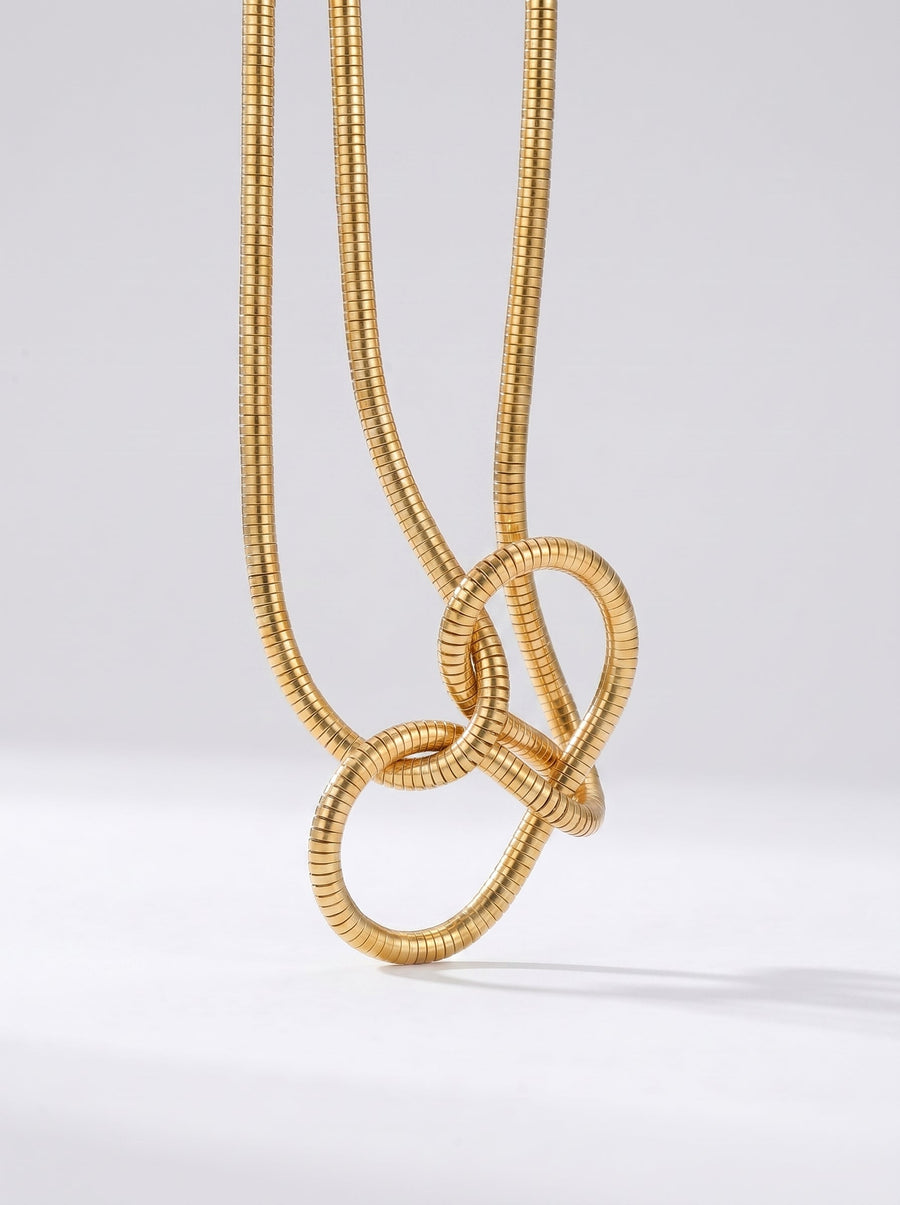 Collar dorado moderno con colgante abstracto en forma de nudo y acabado texturizado, fotografiado sobre fondo claro para un look elegante, artístico y sofisticado.