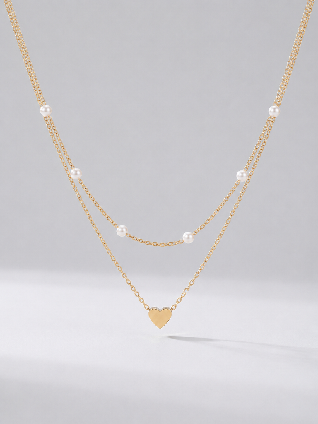 Collar dorado delicado en dos capas con perlas blancas y colgante en forma de corazón, fotografiado sobre fondo claro para un look elegante, romántico y sofisticado.