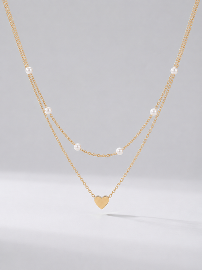 Collar dorado delicado en dos capas con perlas blancas y colgante en forma de corazón, fotografiado sobre fondo claro para un look elegante, romántico y sofisticado.