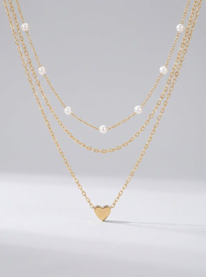 Collar dorado delicado en tres capas con perlas blancas y colgante en forma de corazón, fotografiado sobre fondo claro para un look elegante, romántico y sofisticado.