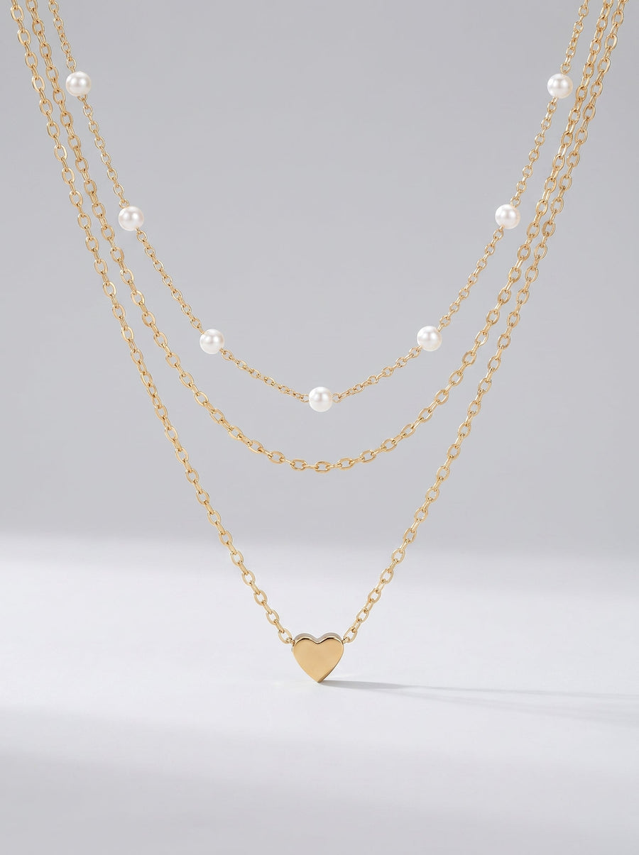 Collar dorado delicado en tres capas con perlas blancas y colgante en forma de corazón, fotografiado sobre fondo claro para un look elegante, romántico y sofisticado.
