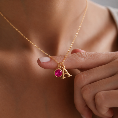 Primer plano de collar dorado con inicial A y piedra de nacimiento rosa, sujeto con la mano para destacar los detalles personalizados.
