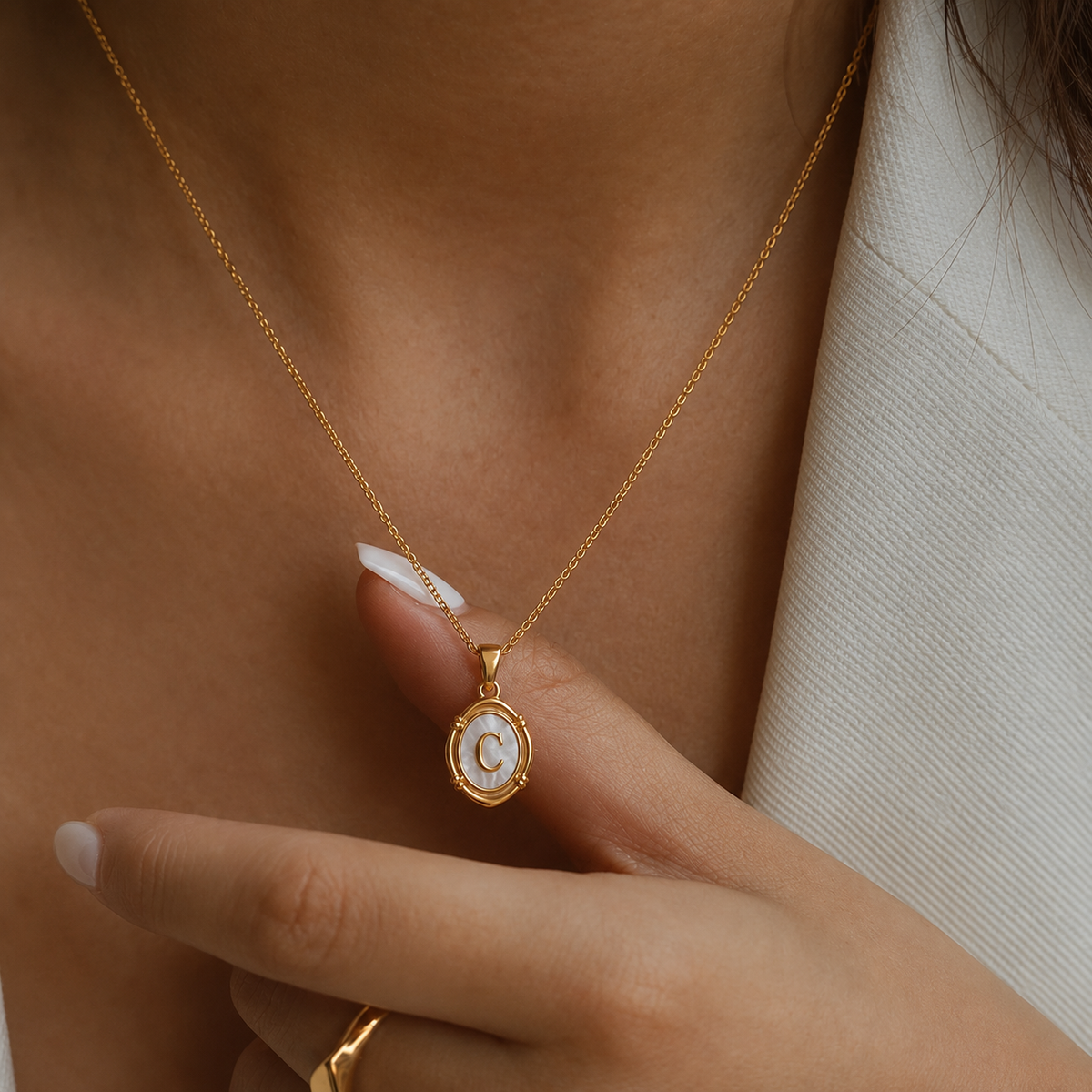 Primer plano de collar oval dorado con inicial C en nácar, llevado al cuello y destacado con la mano.