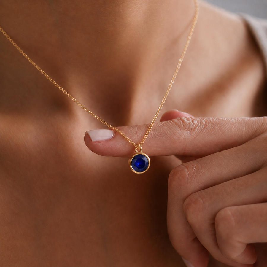 Primer plano de collar dorado con piedra de nacimiento azul (Zafiro), sujeto con el dedo para destacar el colgante circular.
