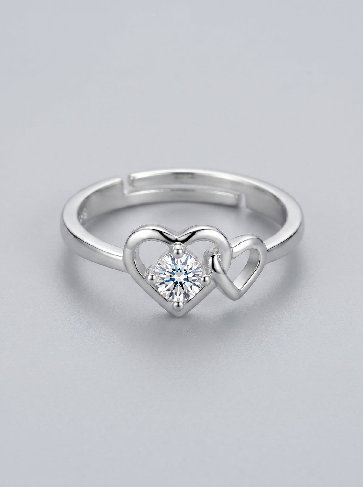 Anillo plateado con diseño en forma de corazón y diamante central sobre fondo gris claro.
