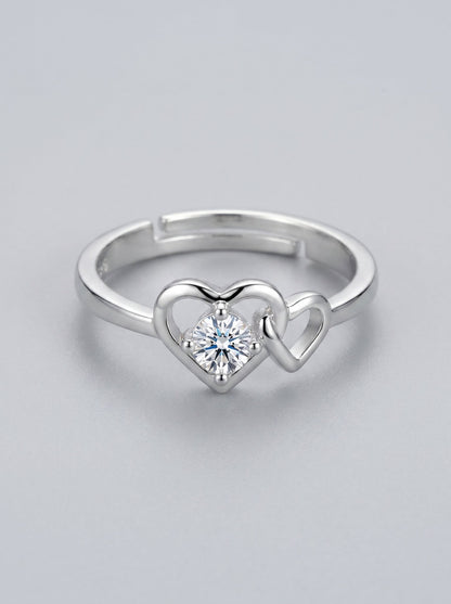 Anillo plateado con diseño en forma de corazón y diamante central sobre fondo gris claro.

