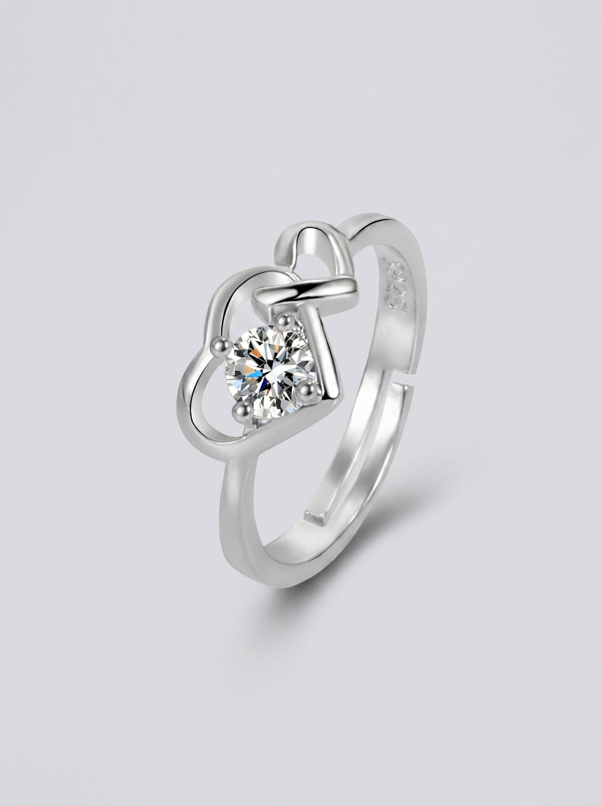 Anillo plateado con un diamante en forma de corazón sobre un fondo gris claro.
