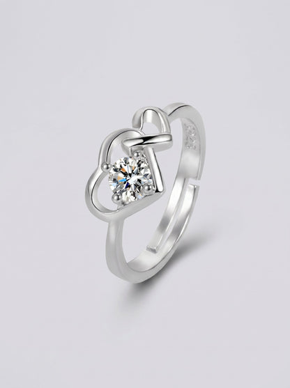 Anillo plateado con un diamante en forma de corazón sobre un fondo gris claro.
