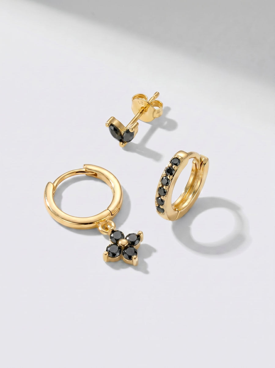 Pendientes de aro y pendientes de botón dorados con detalles en cristal negro, fotografiados sobre un fondo minimalista gris claro.
