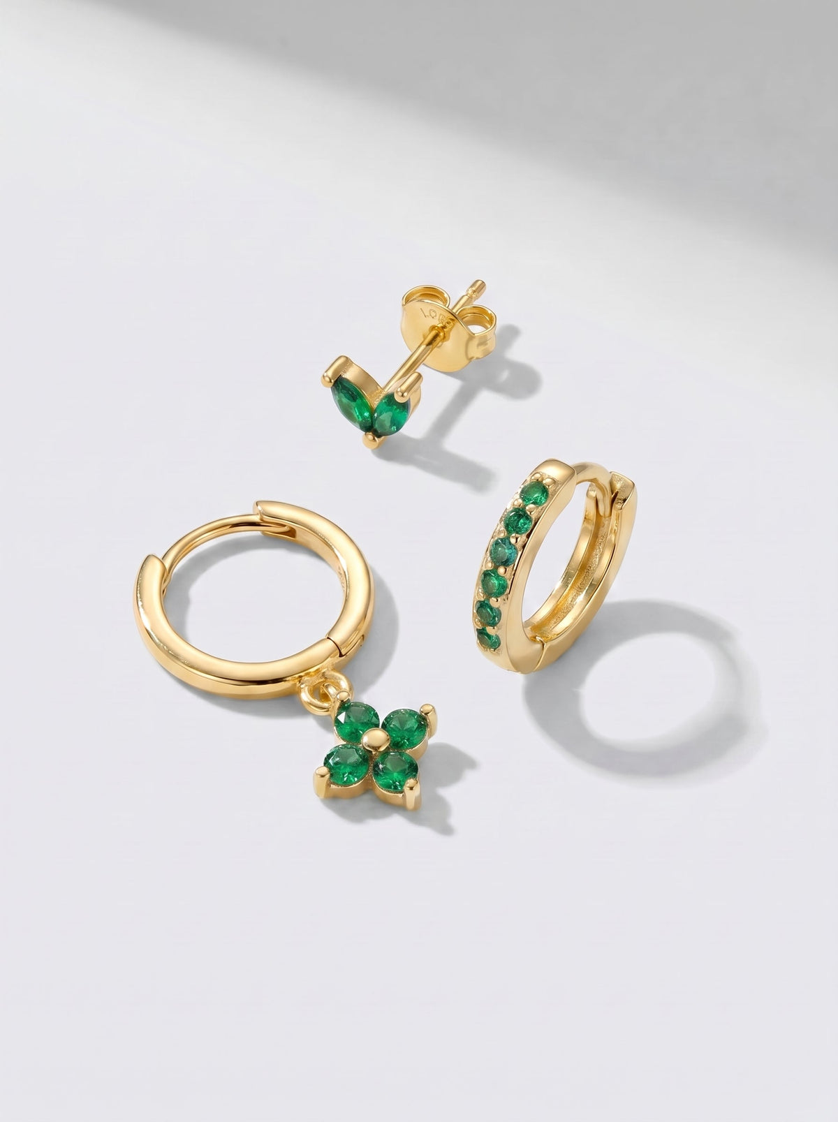Pendientes de aro y pendientes de botón dorados con detalles en cristal verde, fotografiados sobre un fondo minimalista gris claro.
