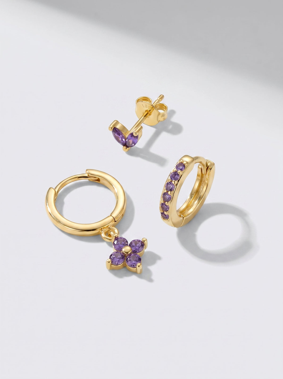 Pendientes de aro y pendientes de botón dorados con detalles en cristal morado, fotografiados sobre un fondo minimalista gris claro.
