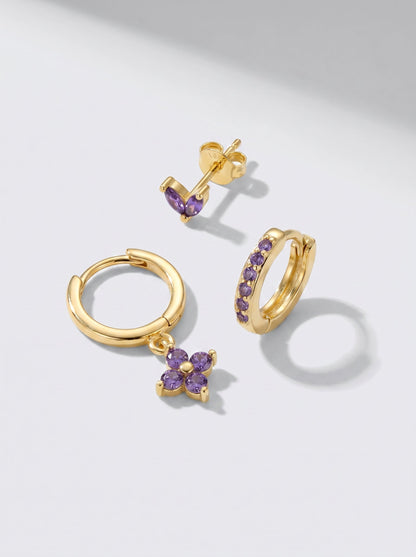 Pendientes de aro y pendientes de botón dorados con detalles en cristal morado, fotografiados sobre un fondo minimalista gris claro.