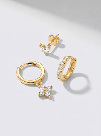 Pendientes de aro y pendientes de botón dorados con detalles en cristal blanco, fotografiados sobre un fondo minimalista gris claro.