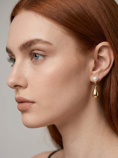 Pendiente dorado colgante con perla blanca y gota metálica, usado por una modelo para un look elegante y sofisticado.