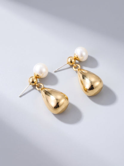 Pendientes dorados colgantes con perla blanca y gota metálica, fotografiados sobre fondo gris claro para un look elegante y sofisticado.
