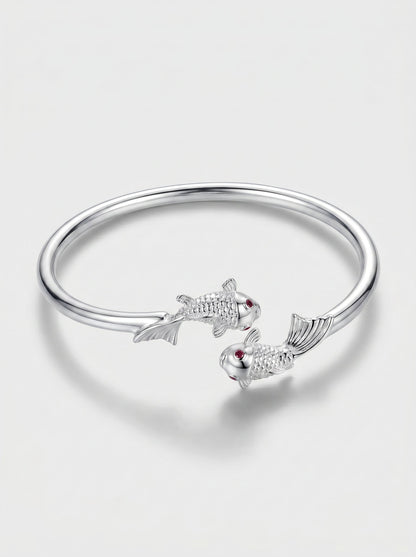 Pulsera plateada rígida con diseño abierto y delicados detalles de peces, fotografiada sobre fondo claro para un look elegante y minimalista.