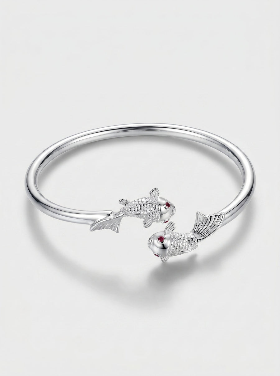 Pulsera plateada rígida con diseño abierto y delicados detalles de peces, fotografiada sobre fondo claro para un look elegante y minimalista.