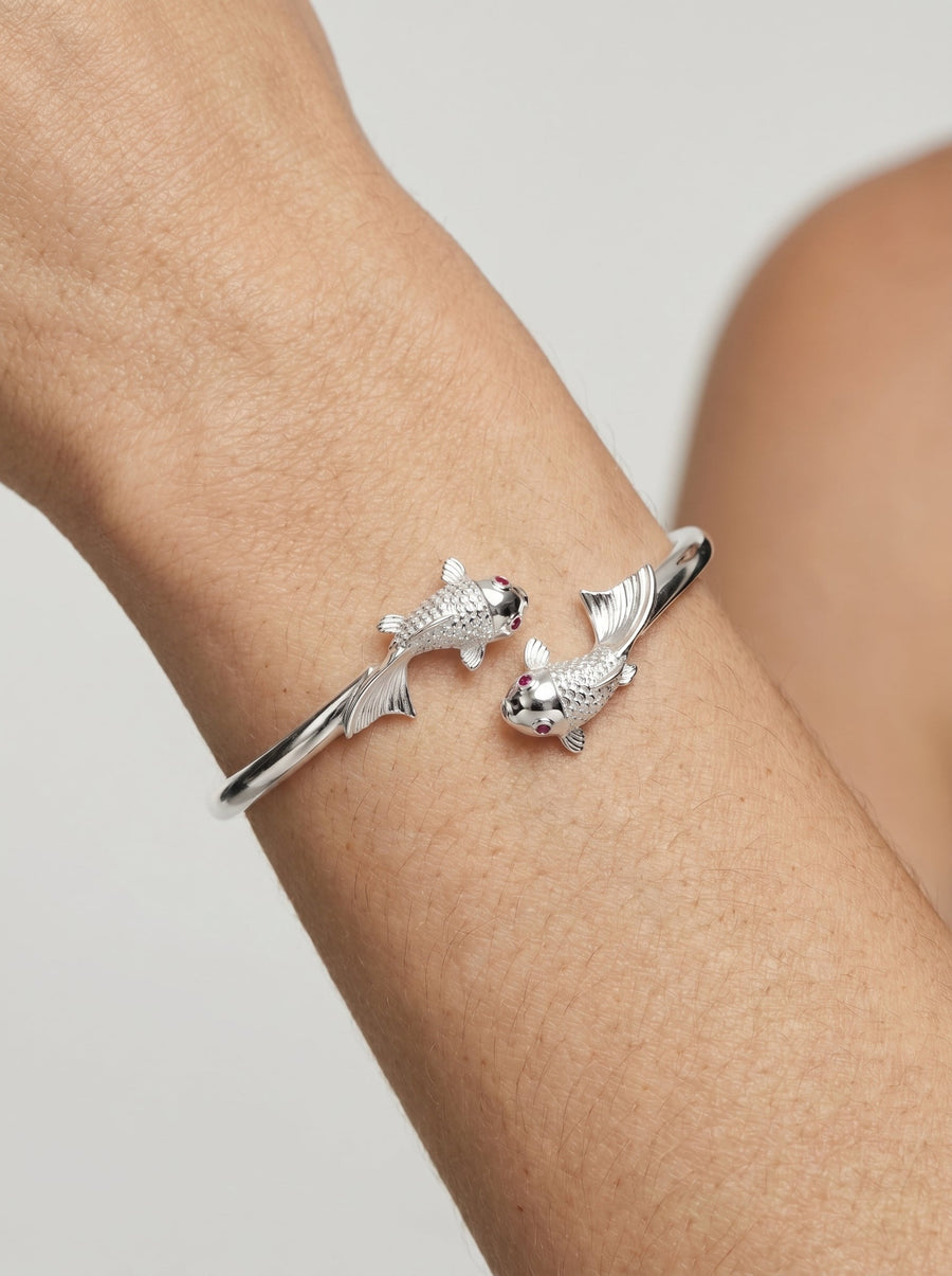 Pulsera plateada rígida con diseño abierto y detalles de peces en los extremos, usada en la muñeca para un look elegante y delicado.