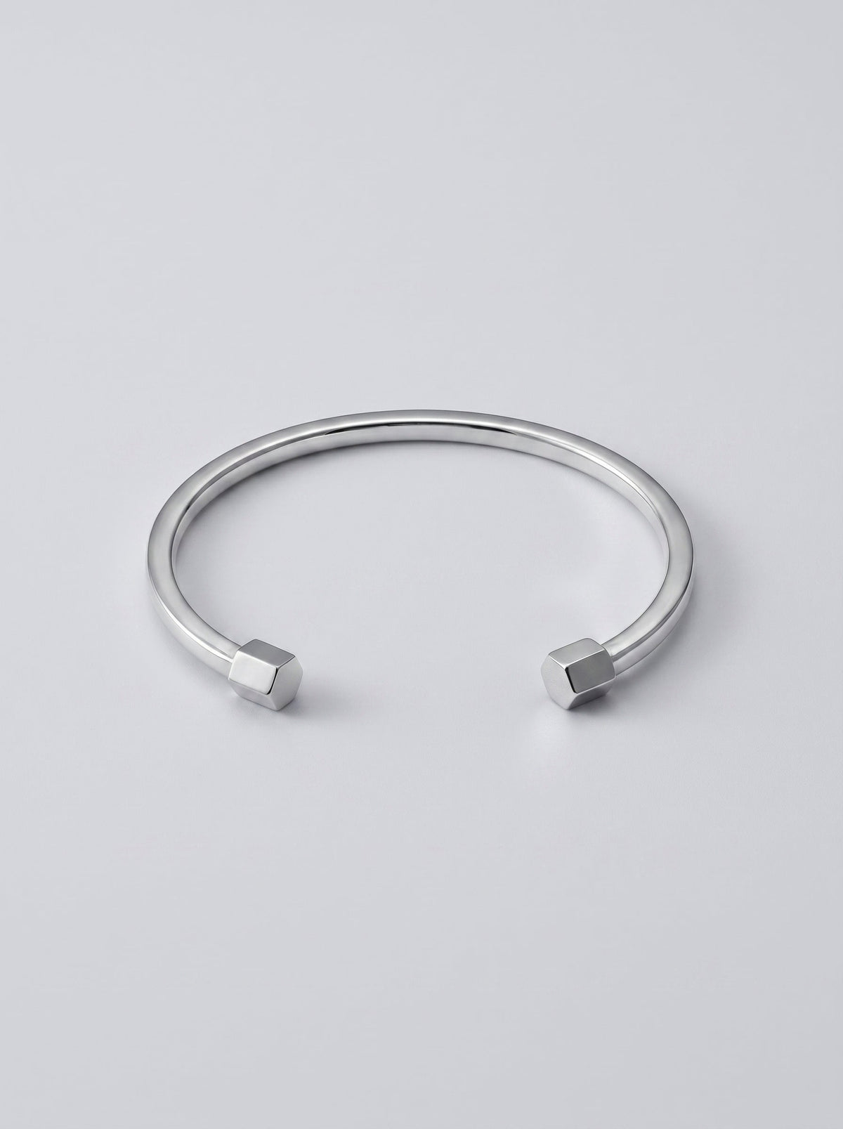 Pulsera plateada rígida con diseño abierto y detalles geométricos en los extremos, fotografiada sobre fondo gris claro para un look minimalista y moderno.