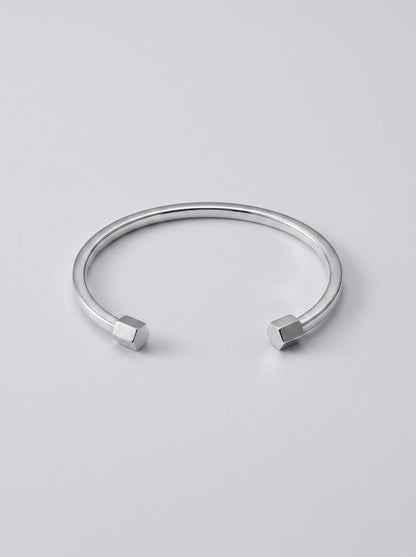 Pulsera plateada rígida con diseño abierto y detalles geométricos en los extremos, fotografiada sobre fondo gris claro para un look minimalista y moderno.