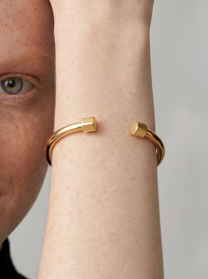 Pulsera dorada rígida con diseño abierto, acabado pulido y extremos hexagonales, usada en la muñeca para un look minimalista y moderno.