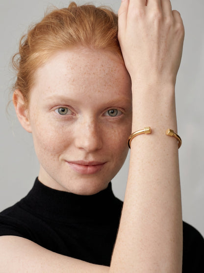 Pulsera dorada rígida con diseño abierto, acabado pulido y extremos hexagonales, usada por una modelo para un look minimalista y moderno.