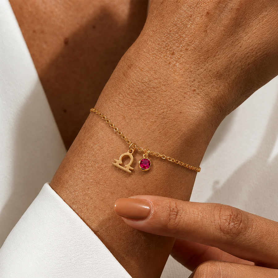 Primer plano de pulsera dorada con símbolo del signo Libra y piedra de nacimiento rosa en la muñeca.