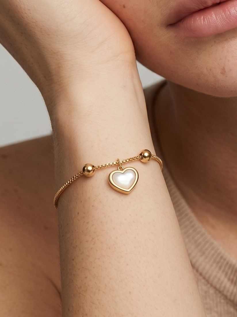 Pulsera dorada delicada con colgante en forma de corazón blanco y acabado brillante, en la muñeca para un look elegante, femenino y romántico.
