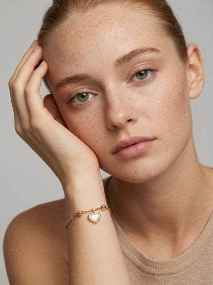 Pulsera dorada delicada con colgante en forma de corazón blanco y acabado brillante, llevada por una modelo para un look elegante, femenino y romántico.