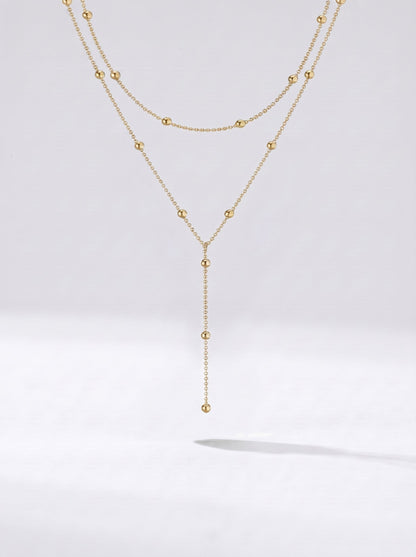 Collar dorado largo en capas con detalles de esferas y colgante delicado, fotografiado sobre fondo claro para un look elegante, sofisticado y moderno.