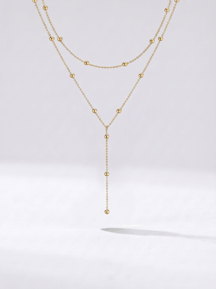 Collar dorado largo en capas con detalles de esferas y colgante delicado, fotografiado sobre fondo claro para un look elegante, sofisticado y moderno.